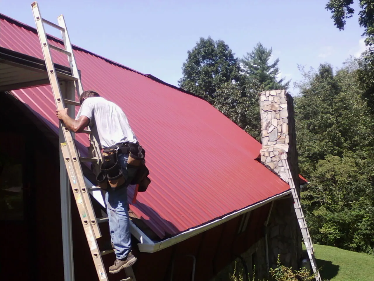 Roofer on a ladder beside a red metal roof and stone chimney — Soffit & Fascia Repair by Roofing Brothers in Weymouth